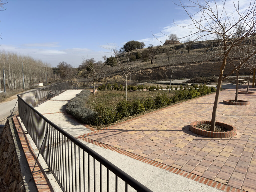 Aprovechamiento del desnivel del terreno a través de una intervención compacta que integra vegetación, caminos accesibles y zonas de reunión, conectando el espacio con su entorno inmediato. La actuación se apoya en la disposición de grandes piedras en seco, conformando muros de contención permeables que permiten la filtración natural del agua y favorecen la respiración del terreno. Esta solución no solo garantiza la durabilidad de la intervención, sino que también refuerza la integración paisajística, potenciando la relación entre arquitectura y naturaleza. El resultado es un espacio público amable, que combina funcionalidad, sostenibilidad y un carácter estético en armonía con el entorno rural.